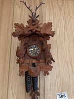Front view of hand-carved wooden cuckoo clock with Roman numeral dial, pinecone weights, and hunting motif including deer head and crossed rifles.