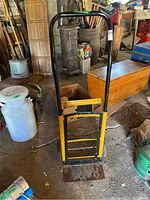 Front view of the yellow and black metal dolly showing the handle, mesh back panel, and base plate.