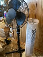 Black adjustable cage fan on stand and white tower 3-speed oscillator fan placed side by side against wood-paneled wall.