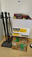 Photo showing black metal fireplace tool set next to cardboard boxes containing fire logs
