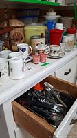 Overview of countertop showing assorted mugs, Tupperware containers, cookie jar, banana hook, rolling pin, and drawer of cutlery below.