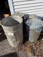 Four metal garbage cans shown outside on pavement next to white siding. One can is open and without lid; three have lids on top. One metal can has a label sticker on the side.