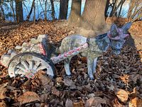 Cement donkey pulling a cart statue on leaf-covered ground with background trees and water.