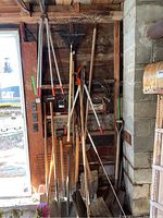 Multiple garden tools lined up inside a wooden shed. Visible are shovels, a metal rake with a red handle, pruning shears with orange handles, and various long-handled tools leaning against the wall.