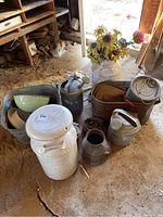 Overview of multiple galvanized and enamelware items including milk cans, watering cans, tubs, bowls and horseshoes on dirt floor