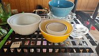 Lot of Pyrex and Fire King mixing bowls in various colors and sizes on a metal shelf, also includes glass baking dishes and a pottery bowl.