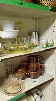 Shelf with assorted glassware including yellow-handled glass caddy with six shot glasses, brown ceramic mugs, egg cups, glass cups, and wooden containers.