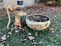 Wide shot of the hand pump and round metal pot outdoors on grass with fallen leaves