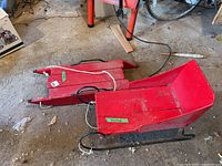 Photo of two vintage wood items: a red wooden sleigh and a wooden sled with rope attached, placed on a rough cement floor.