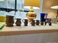 Photo of eight antique pewter miniature vessels arranged in a line on a white table cover with labeled lot number. Includes various sizes of cups and pitchers with dark pewter patina.