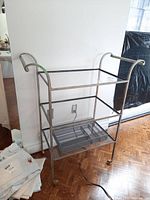 Metal frame cart with 3 glass shelves and a metal cutlery organizer tray on bottom shelf, on wheels, shown at an angle.