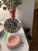 Photo of a round pottery plate with detailed cherub and floral motif, pink glazed ashtray, and part of a clear crystal vase with dried eucalyptus
