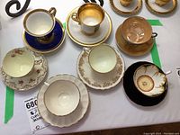 Wide top-down view showing all seven cups and saucers, each with distinct colors and trim finishes, arranged on a white surface.