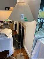 Floor lamp with beige cone-shaped fabric shade, tall metallic stand, and square base on wood floor.