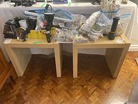 Overview photo showing the lot items arranged on two small tables including flashlight, extension cords, hardware pieces, lamps, steamer, and power bars