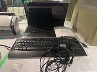 Open Toshiba Satellite laptop with black Microsoft wireless keyboard on the keyboard area, charger cables loosely coiled on the keyboard.
