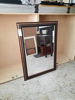 Full view of the wood framed wall bevelled mirror, showing the entire mirror standing against a shelving unit on a tiled floor.