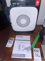 Front view of NOMA oscillating ceramic heater showing the white front grill with circular vent pattern, instruction manual, lot tag, and packaging box.