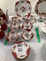 Photo shows six teacups and matching saucers stacked along with six dessert plates, cake plate in back and milk jug, all in Paragon Rockingham pattern with floral and red/gold design.