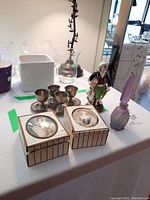 Photo showing miniature pewter cups on tray, porcelain figurine, frosted glass perfume bottle, and two decorative boxes with portraits on lids