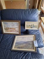 Three framed watercolour prints resting on a dark blue leather surface. One larger framed print shows wooden boats by water, another medium-size frame with a landscape of hills or mountains, and a smaller framed print with a rural scene featuring a stone bridge and trees.