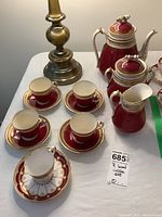 View of overall coffee set with four matching cups and saucers, one additional non-matching cup and saucer, and three coffee/tea pots on white tablecloth beside brass lamp base.