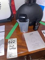 Close-up of black round Saje aroma diffuser with included measuring cup and bottle of essential oil scent on wooden surface.
