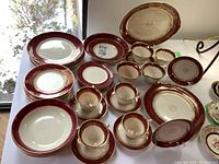Full view of the china set laid out on a table by a window, showing the burgundy and gold pattern on white porcelain. Includes all plates, bowls, cups, and saucers described by the seller.