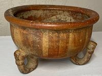 Front view of the pottery vessel showing round bowl shape and three curled tripod legs with surface wear.