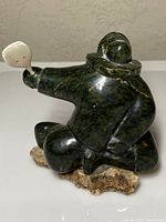 Front three-quarter view of dark green mottled soapstone figure kneeling with hooded parka, holding white bone implement