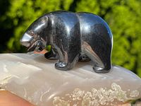 Side view of black polished hematite bear sculpture on translucent stone base with natural crystal inclusions, outdoors in natural light.
