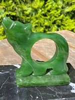 Green jade bear sculpture with circular body cutout sides and black marble base shown outdoors on wooden surface with natural light highlighting texture and color of jade.