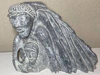 Front and angled perspective of large Inuit soapstone sculpture showing main figure's face, headdress, and smaller face carved at base within opening.