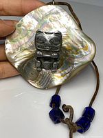 Front view of square abalone shell pendant with carved metal totemic figure and leather cord.