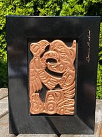 Front view of carved copper panel mounted on a black frame, showing Northwest Coast design and Clarence Mills signature.