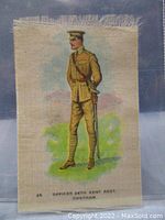 Full view of an antique silk textile depicting an officer of the 24th Kent Regiment in uniform, standing on green ground with a blue sky background. Approximate size 3 by 2 inches, with frayed edges visible.