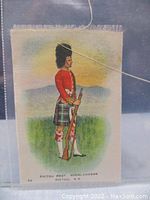 Photo of an antique tobacco silk measuring approximately 3 by 2 inches showing a colored print of a Pictou Regiment Highlander soldier in red jacket, kilt, and fur hat with a rifle, standing against a scenic background.
