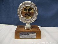 Front view of the trophy with wooden base and silver top showing tennis rackets emblem