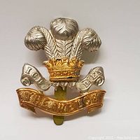 Front view close-up of the bi-metal Welch Regiment cap badge showing silver feathers and crown with gold accents.
