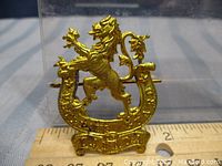 Front view of the Veteran Guards of Canada gold-tone metal cap badge on a ruler for scale, showing detailed lion and horseshoe design with banner below.