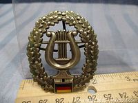 Front view of metal beret badge with lyre surrounded by laurel wreath and West German flag emblem at base, ruler shown for scale.