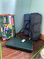Green Coleman two-burner propane stove closed next to two blue tents packed in bags and orange shovel