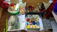 Photo showing several boxes containing assorted Christmas ornaments including baubles in different colors and sizes, a decorative stone heart, gold leaf-shaped ornaments, and folded winter-themed green fabric.