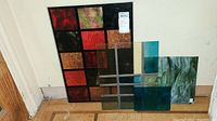 Four multicolor stained glass panels leaning against the wall on a wooden floor showing various colors including red, black, orange, green, teal, gold, and marbled green.