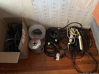 View of a collection of coiled white and black cables on floor plus power strips and plugs tangled on the side. Also shows several cable rolls including a red spool.