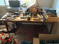 View of wood dining table covered with an assortment of hand tools and hardware parts including clamps, drills, measuring tapes, and power tools.