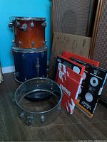 Photo showing two tom drums stacked, one brown and one blue, with several boxed drumheads and a metal drum frame visible.