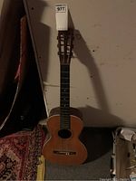 Front full view of the brown six-string acoustic guitar standing upright against a wall, showing the guitar body, sound hole, fretboard, and headstock.