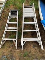 Top view of two aluminum ladders laid on grass, showing one with fold-out black plastic tray shelf and one standard ladder
