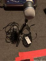 Photo showing the large silver-gray microphone mounted on a small tripod microphone stand, one smaller handheld microphone with a coiled cable, one slim black microphone with XLR connector, and several cables.